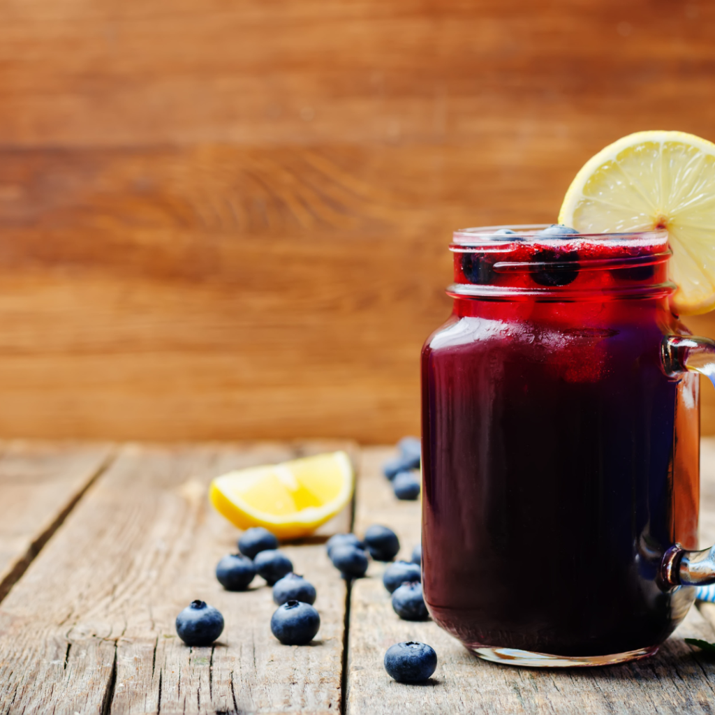 "blueberry lemonade in glass jar with ice and a lemon slice with blueberry and lemon wedges on a wood background"