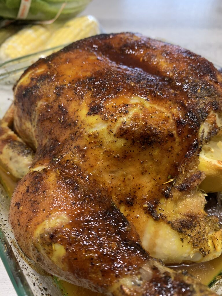 "A Golden brown roasted whole chicken in clear dish with corn on the cob in the background"
