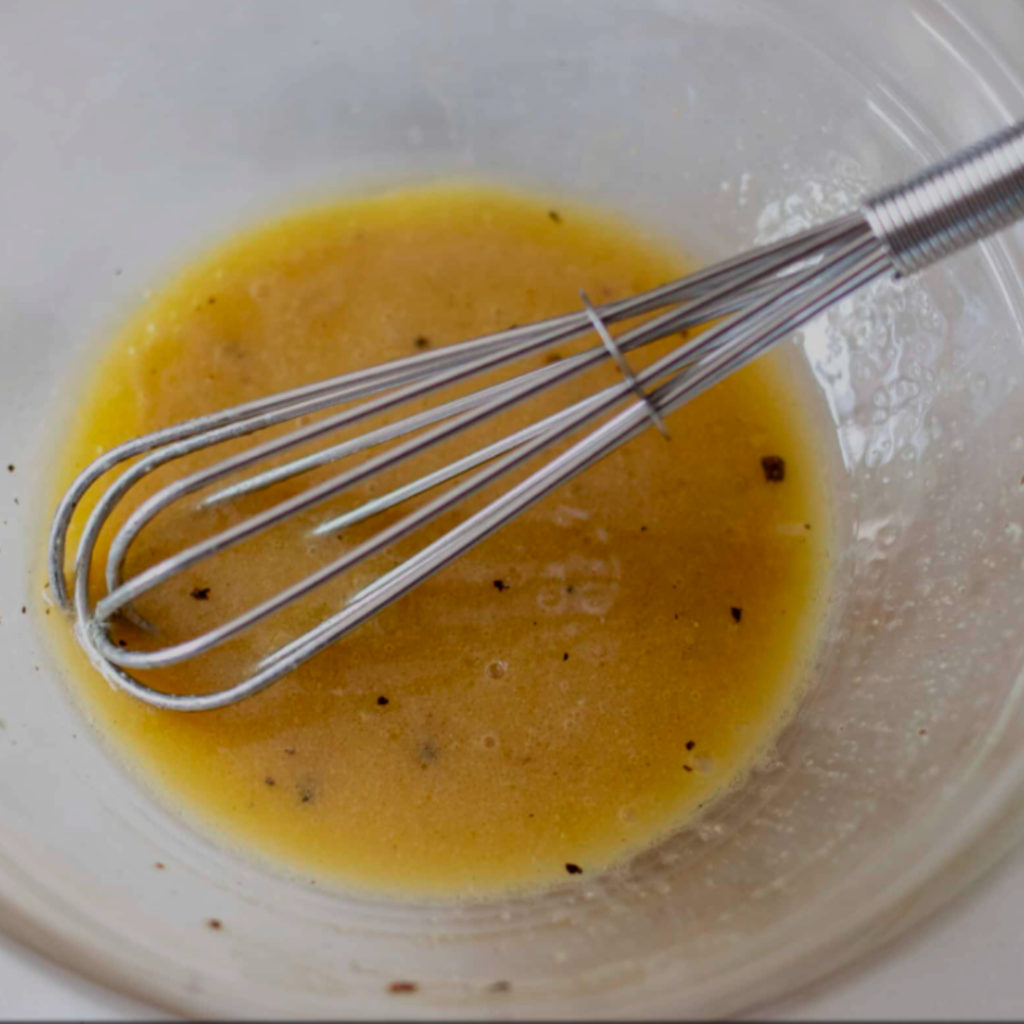 "Dijon Vinaigrette with a whisk in a glass bowl on a white tile countertop"