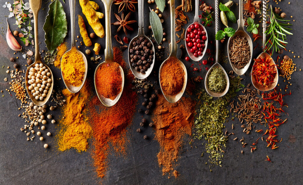 "Nine global spices in hues of red, yellow, brown and green in silver spoons on black background"