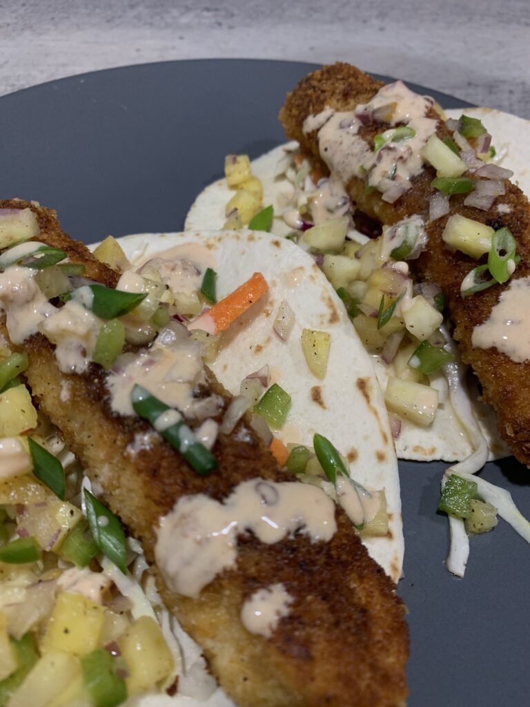 "breaded fried tilapia, with carrots, green onions, white sauce, taco shell on a gray plate"