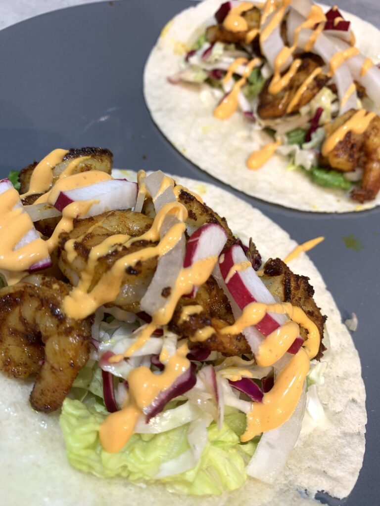 "fried shrimp, with a spicy sauce, lettuce, radishes, red onions on a taco on a gray plate
