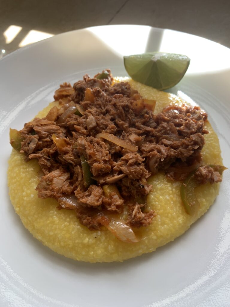 "Canned tuna with onions, tomatoes, bell pepper, tomato paste on yellow grits on a white plate with a wedge of lime"