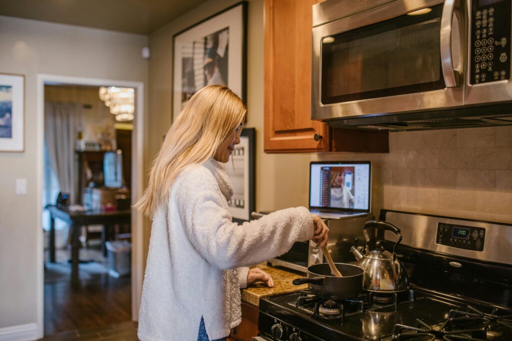 "White lady with blond hair at the stove stirring a pot with wooden spoon while watch some on a computer laptop"