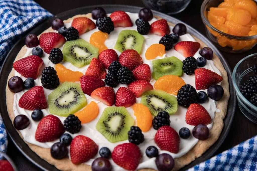 "red strawberry, kiwi slices, whole blueberries. orange segments on cream cheese frosting and crust to create a fruit pizza