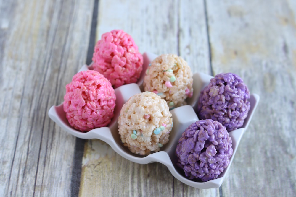 "Pink, purple, neutral easter egg shaped rice krispies treats in a egg carton on a wooden table