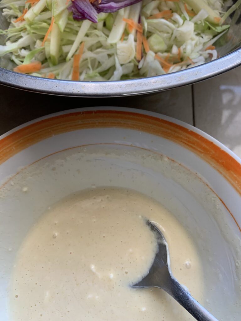 "coleslaw dressing in a bowl with an orange trim with coleslaw ingredients in the background"