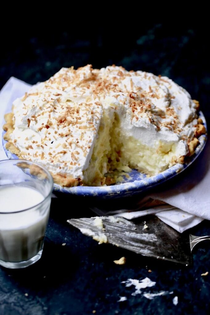 "A pie crust filled with a layer of coconut custard, meringue topping and toasted coconut top. With a glass of milk on a black counter top