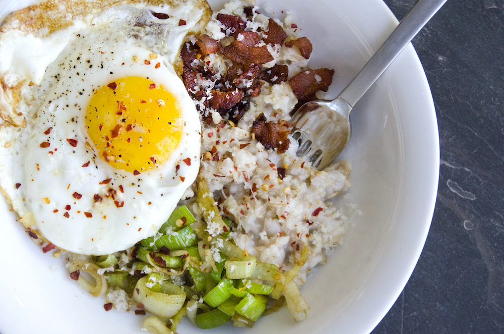 "Oats with crumbled bacon, egg and green onions on a dark countertop its in a white bowl and a has a silver fork on the side