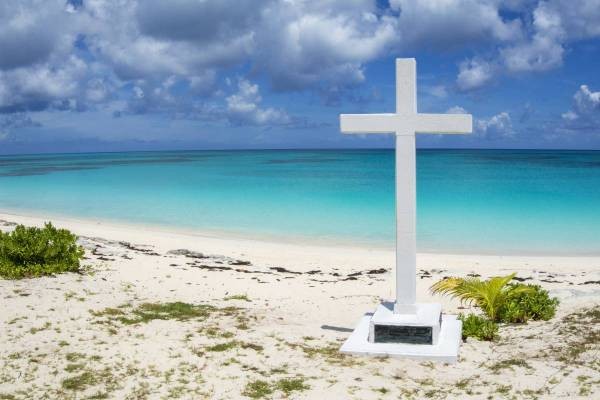 " A white cross monument on a white sandy beach with the ocean in the background