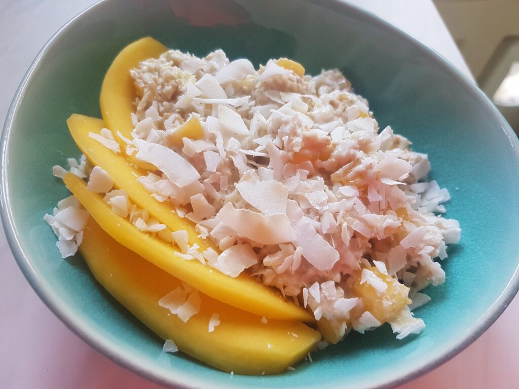 "Overnight oats with slices of mango and coconut in a blue green bowl o a white background