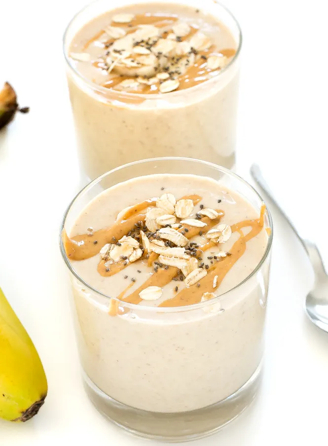 "Clear glasses that have oatmeal smoothies made with banana and oats, its on a white background with a spoon and a banana in the background"