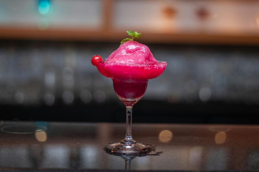 "A deep pink drink with a green herb sprig on top on a glass bar top with glasses in the background"