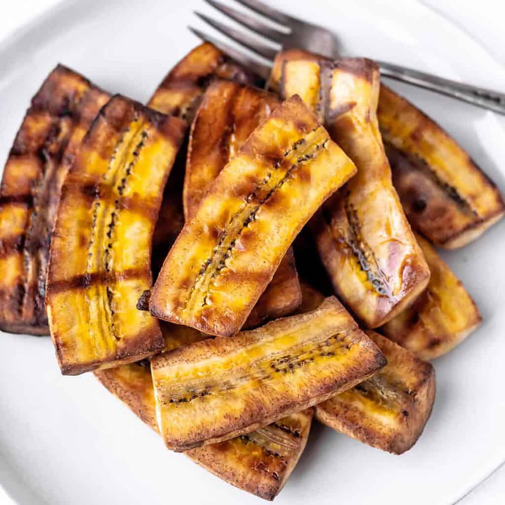 "Grilled cooked plantain on a white plate with a fork"