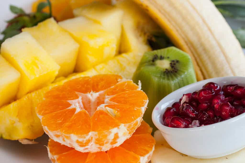 "fruits on a counter orange, banana, pomegranate seeds and pineapple