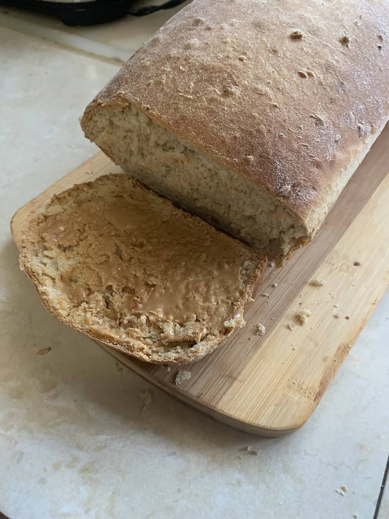 A loaf of bread with slice of bread covered in peanut butter on a cutting board