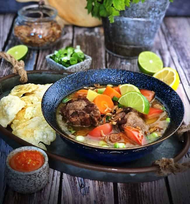 A black bowl filled with oxtail meat, vegetables and broth with a lime wedge, with naan bread and condiments on the table