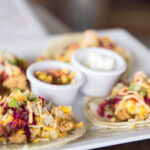 "4 tacos on a white plate with a salsa and sour cream"